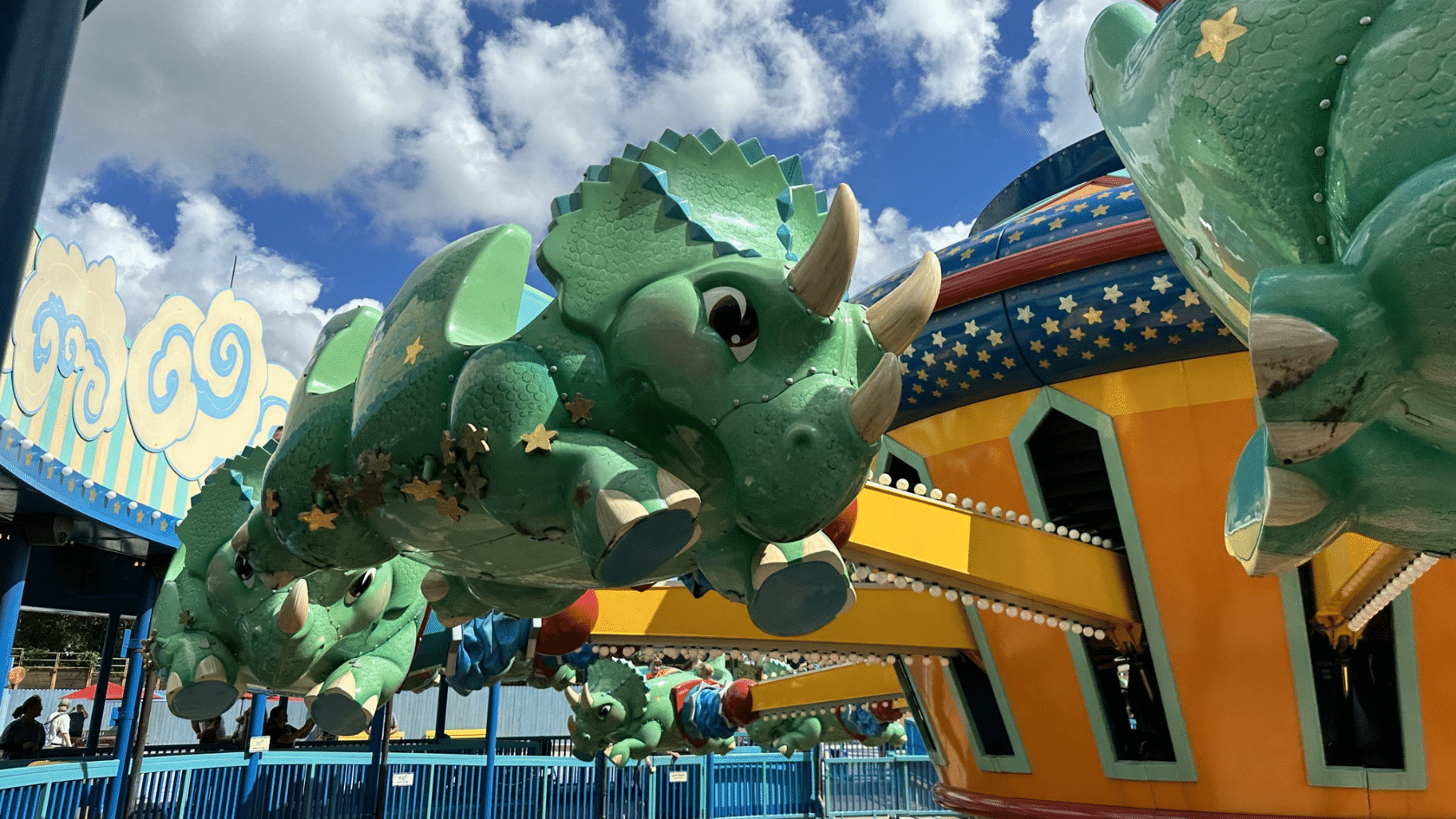 green triceratops figures circling on a carnival ride beneath a bright blue sky filled with fluffy clouds