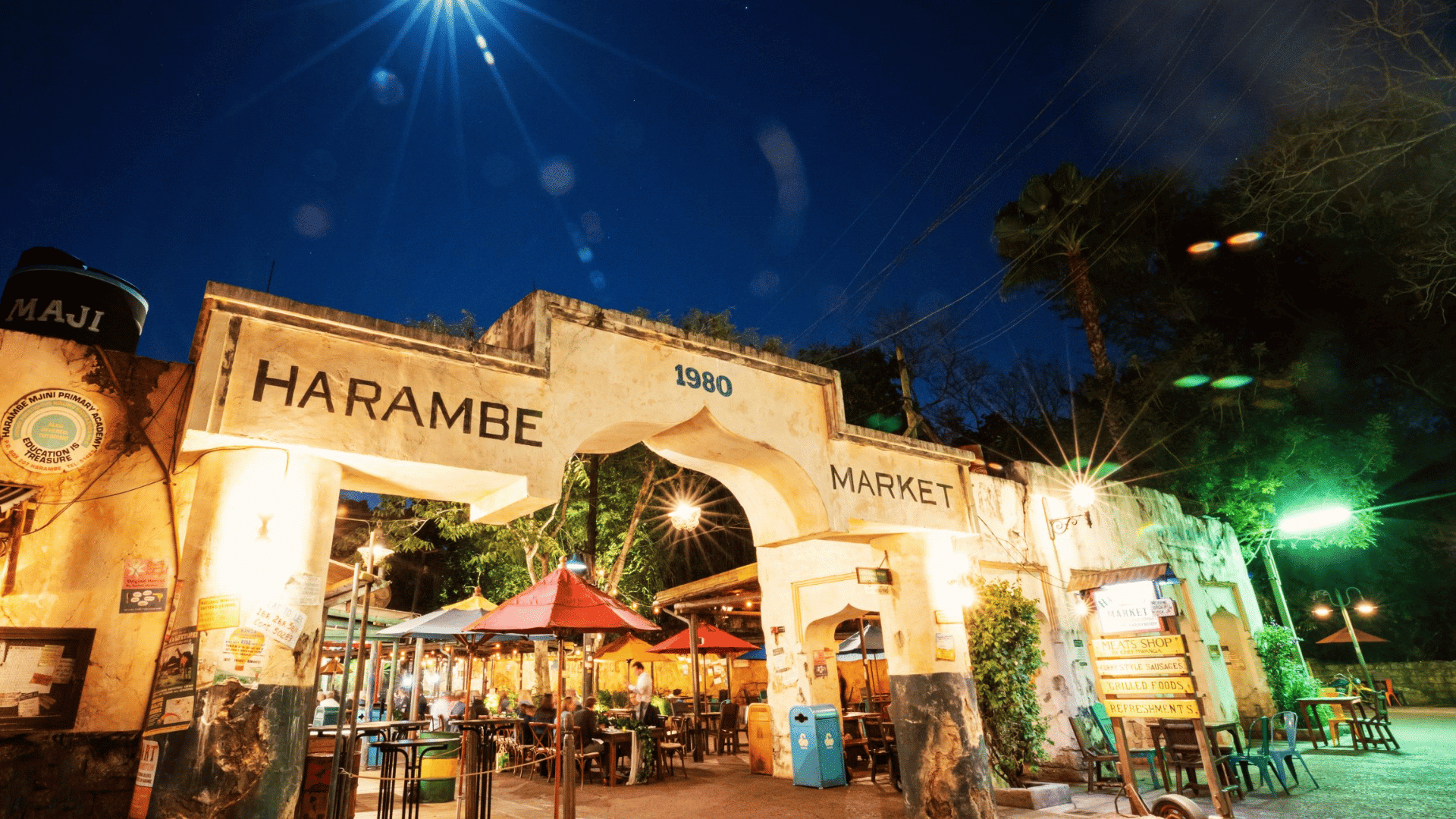 harambe market entrance illuminated at night with outdoor dining tables colorful umbrellas and a bright moon overhead