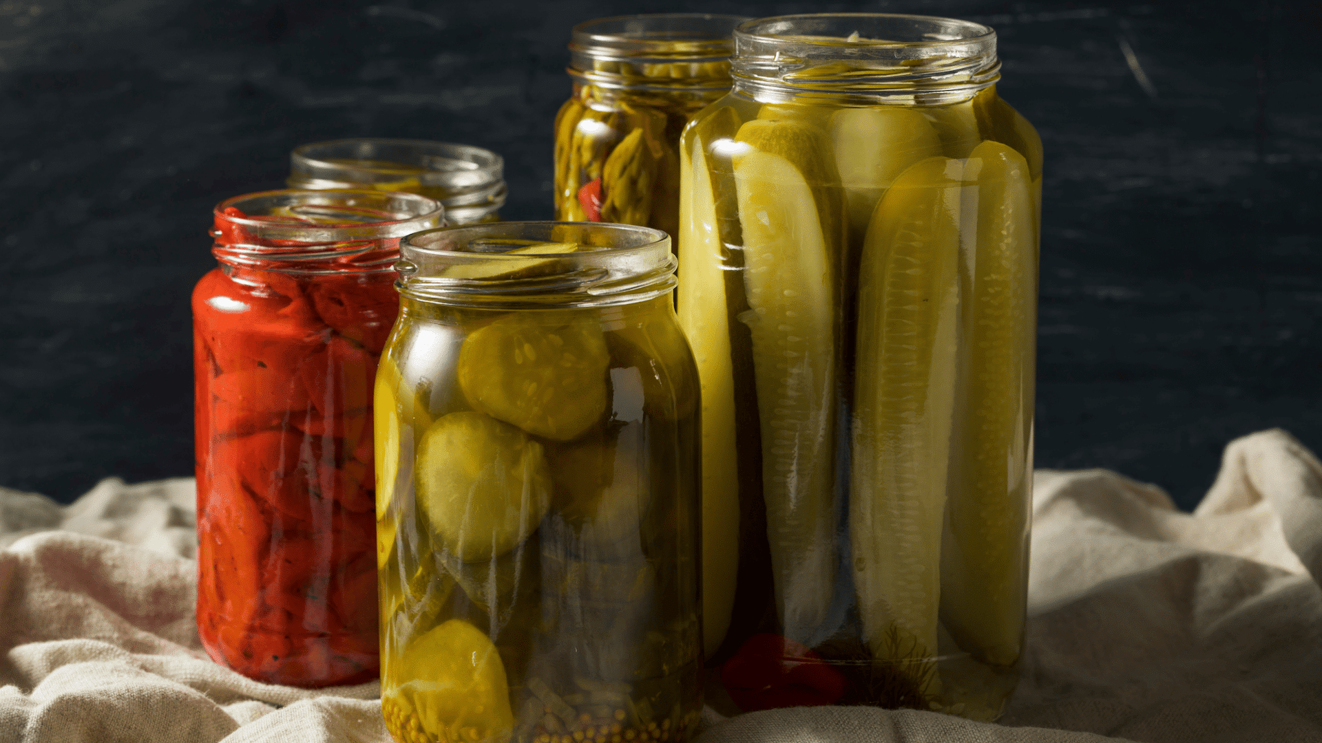 jars of pickled cucumbers and red peppers on cloth against a dark kitchen background
