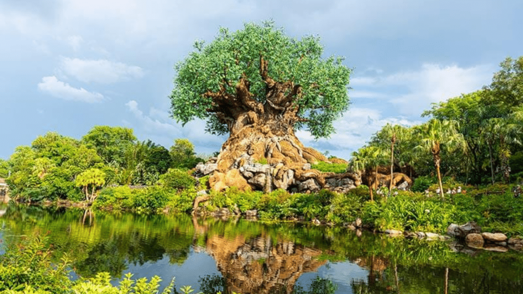 massive tree with sprawling roots beside a calm pond surrounded by lush greenery beneath a cloudy sky reflected in water