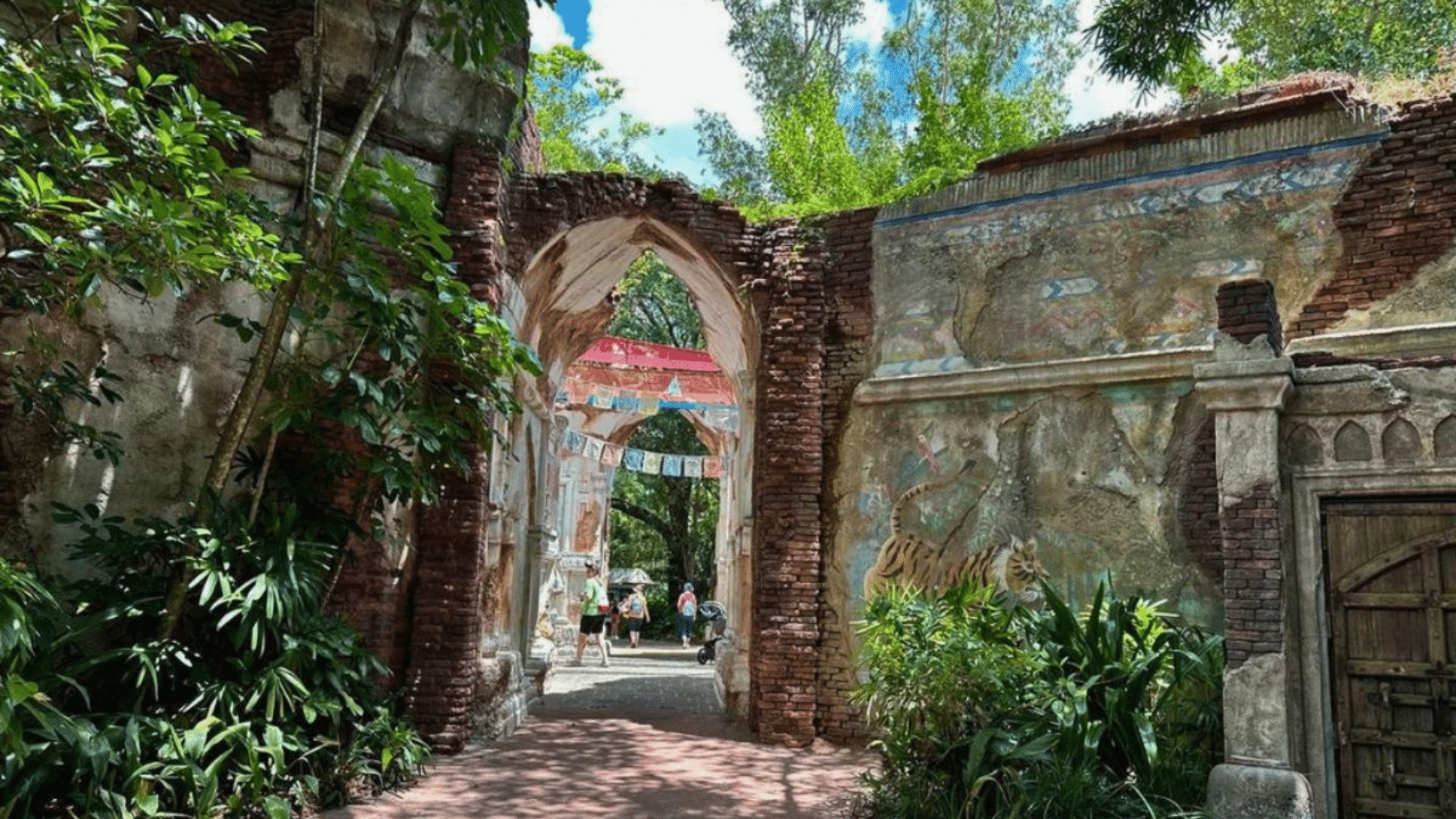 pathway passes through a weathered stone archway with faded murals lush plants and visitors walking beyond the entrance