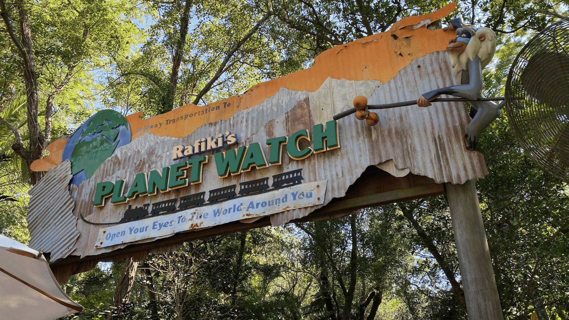 rafikis planet watch entrance sign stands among lush trees with rafiki character figure and railway themed details above path