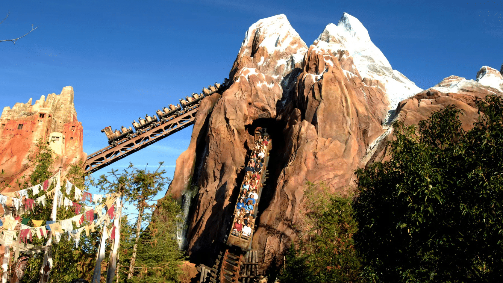 roller coaster train carrying riders descends through a rocky snowcapped mountain surrounded by trees and blue sky