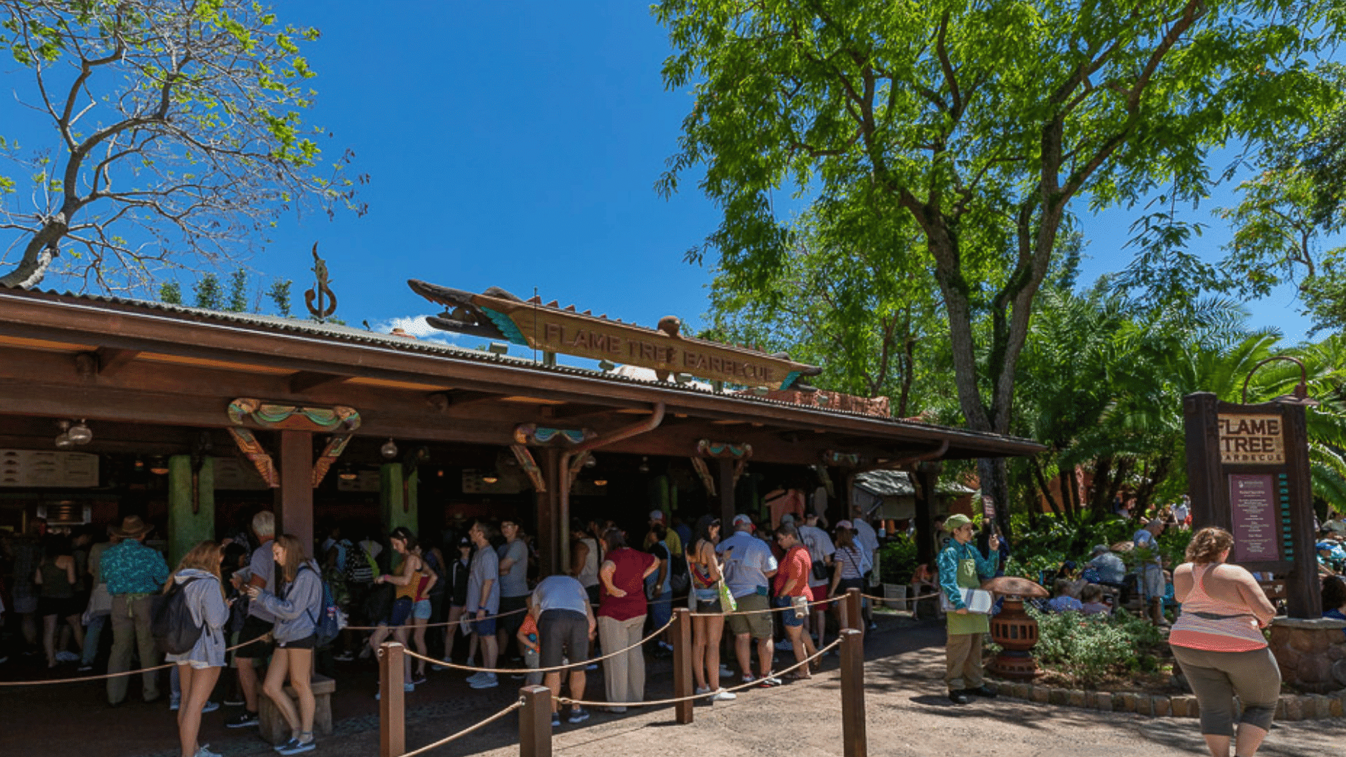 visitors wait in line outside flame tree barbecue beneath tall trees on a sunny day in a busy theme park