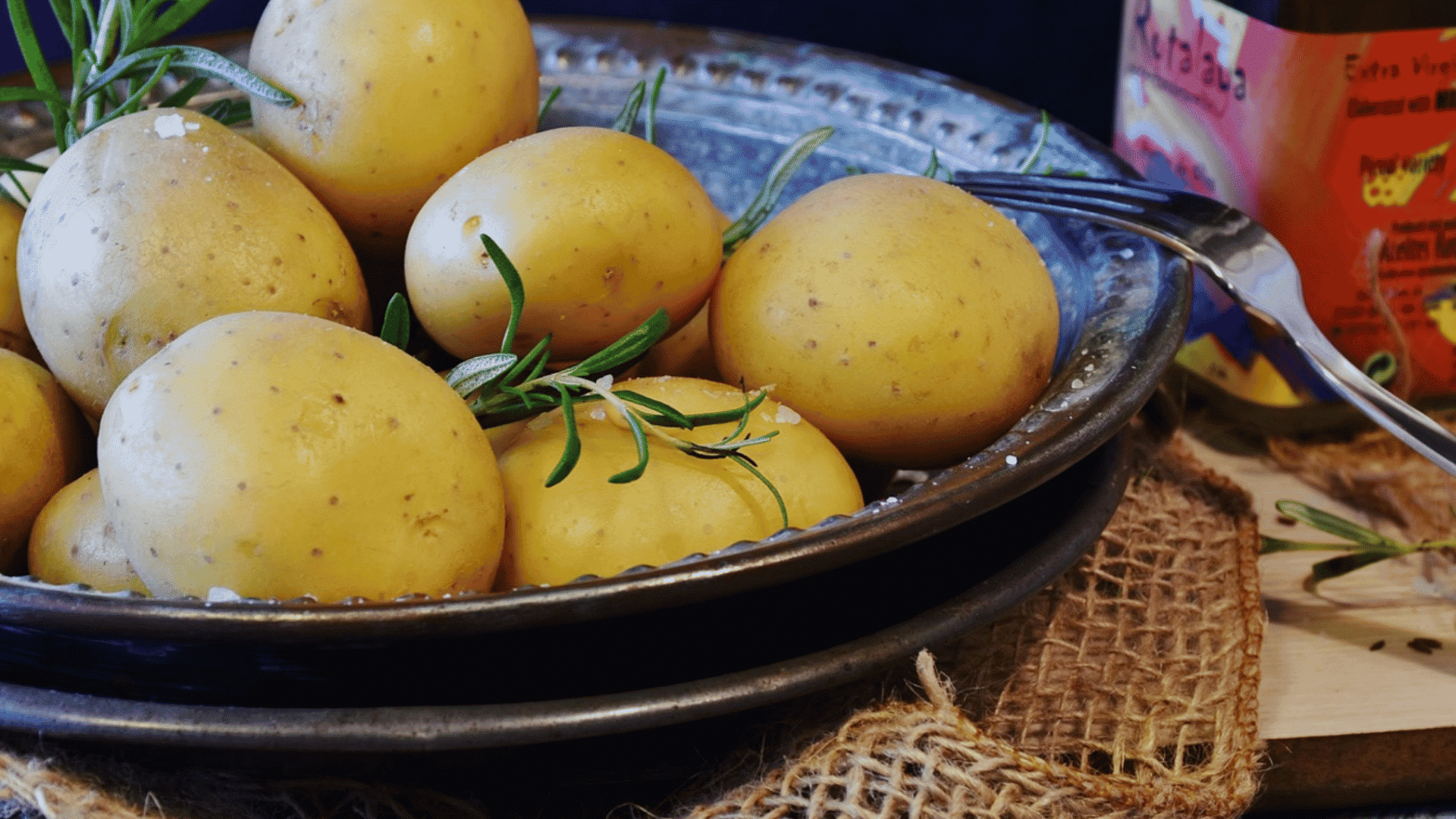 yellow potatoes with rosemary and coarse salt sit in a metal dish beside a fork and bottle of olive oil
